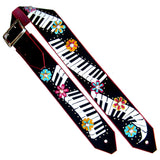 A black guitar strap featuring a white piano keyboard pattern and colorful floral motifs, with a dark red leather trim and a silver metal buckle. Owned by Kristin Chenweth.